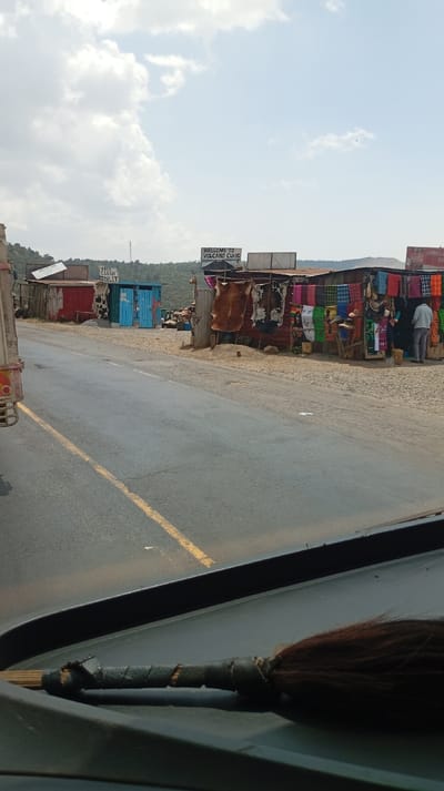 The image depicts a roadside market scene located in Lari/Kirenya ward, Kenya, viewed from inside a vehicle. A two-lane asphalt road with a solid yellow dividing line extends from the bottom center into the midground. On the left, the rear section of a large, red and white truck is partially visible. The foreground shows a dark-colored vehicle dashboard with a wooden stick featuring a dark, fibrous tassel (possibly a fly-whisk) resting horizontally.
Along the right side of the road, a series of makeshift market stalls are constructed from corrugated metal and painted wood. The closest prominent stall on the right features a sign reading "WELCOME TO VOLCANO CURIO S." This stall displays multiple animal hides, including a large brown one and a black-and-white spotted one, along with numerous colorful textiles and woven goods. One individual wearing light-colored trousers and a shirt stands within this stall, facing away from the camera. Further right, a partial view of another stall shows a "Coca-Cola" sign and displays more colorful fabrics.
Further down the road, on the right, is a distinct blue structure with a sign proclaiming "FREE CLEAN TOILET." Behind this, another red-painted stall is visible. The ground immediately adjacent to the road and around the stalls is unpaved, appearing dry and dusty. In the background, green hills with sparse vegetation rise under a sky with scattered white clouds. The overall scene suggests a commercial stop along a rural or semi-urban roadway.