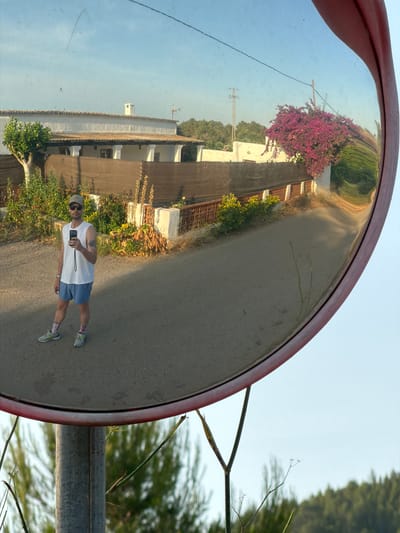 In Santa Eulària des Riu, Spain, a man is pictured reflected in a convex mirror mounted on a metal pole. The man, wearing a white tank top, blue shorts, a baseball cap, sunglasses, and athletic shoes with colorful socks, holds a smartphone in his right hand, appearing to take a selfie. He has tattoos on his left arm. The mirror's reflection shows a rural road bordered by vegetation and a low fence. In the background, a white house with a tiled roof is visible, along with a vibrant pink bougainvillea bush. The sky is clear and light blue. The scene is captured during daylight.