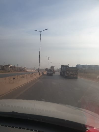 The image captures a roadside scene, likely from the perspective of a passenger in a moving vehicle, looking forward and slightly to the left. The setting is a wide, multi-lane road or highway in Killi, Syria, under an overcast and hazy sky.
In the foreground on the left, a tall, black street light with a distinctive Y-shaped lamp fixture stands prominently against the dull sky. Further down the road, another smaller street light is visible. The road itself is made of asphalt, with a lighter-colored shoulder or barrier separating it from the unpaved or dirt area on the left. This area features what appear to be piles of construction materials or rubble, and some distant, indistinct buildings, suggesting ongoing development or an industrial zone.
On the main road, several vehicles are present. A large, dark-colored truck, possibly a dump truck or flatbed, is visible in the middle distance, traveling in the same direction. Other smaller vehicles can be seen further ahead on the road.
The right side of the frame is occupied by a portion of the interior of the vehicle from which the photo was taken, including part of the windshield pillar and possibly the dashboard, which are light in color. The overall impression is one of travel on a busy road during a daytime with subdued light due to the heavy cloud cover. No specific human activity is discernible, nor is any text visible.