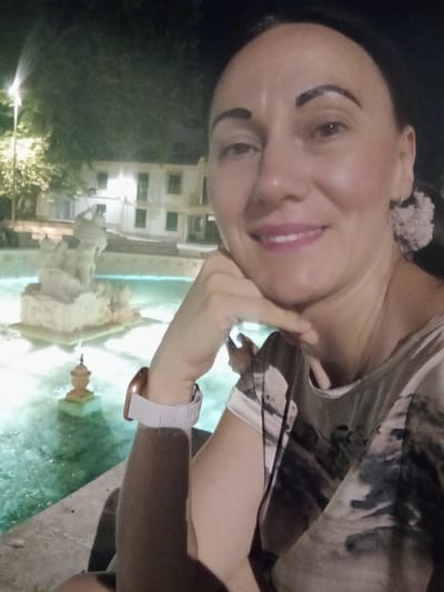 A woman is captured in a selfie at night, with a fountain and illuminated buildings in the background. The fountain features a sculpted element and is filled with turquoise water, with a smaller decorative urn visible within it. The buildings, partially obscured by trees, are lit, suggesting an urban setting. The woman is wearing a light-colored shirt with a patterned design and a white smartwatch on her left wrist. Her right hand is resting on her chin, and she is looking towards the camera with a slight smile. The overall lighting suggests it is evening or night. The background architecture and fountain are characteristic of the Fuente de la'$.