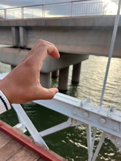 A hand is positioned in front of a bridge structure with water visible in the background. The bridge is constructed of concrete and steel, including white support beams secured with bolts. A wooden plank is located near the edge of the image. The hand is light-skinned and partially obscuring the view. The location appears to be outdoors. Without additional information, the precise location within Tabaroucht, Morocco, cannot be determined.
