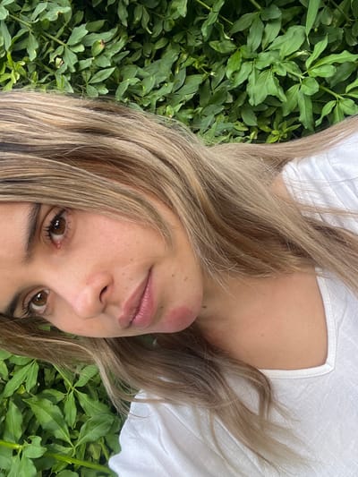 A fair-skinned woman with blonde hair lies in green foliage. She wears a white top. Her face is tilted, and she looks directly at the camera. The background is filled with lush green plants, creating a natural setting. Her lips are light pink. Her eyes are brown.