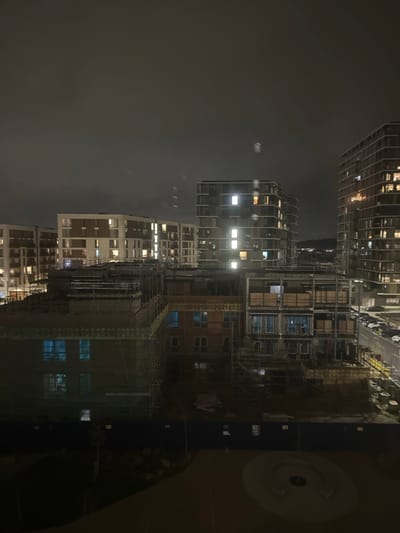 The image depicts a nighttime urban landscape in Gillingham, United Kingdom, specifically aligning with regeneration areas such as the Gillingham Waterfront or developments near the Medway Campus, characterized by modern multi-story residential buildings and active construction.
The scene is dominated by multiple residential structures under a dark, overcast sky. In the background, several completed multi-story apartment blocks are visible; those on the left feature light-colored facades with darker contrasting panels, while the taller building on the far right presents a darker, possibly glazed, exterior. Numerous windows across these completed buildings are illuminated, indicating occupancy.
In the midground, an extensive construction site is evident, featuring partially built multi-story structures. These buildings display exposed brickwork and are extensively covered with scaffolding. Some windows within these incomplete structures show a distinct blue glow, possibly from internal lighting or protective coverings. The ground within the construction zone appears unpaved and uneven.
The foreground reveals a dark, open communal or park area, separated from the construction site by a dark blue hoarding fence. A circular paved feature, possibly a seating area or decorative element, is discernible in this park area. On the far right, a street with multiple parked vehicles is visible adjacent to the construction site. No people, specific actions, or interactions are discernible in the scene. The lighting is exclusively artificial from building interiors and streetlights.