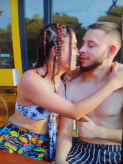 In Hrishteni, Bulgaria, a couple embraces closely, their faces nearly touching. The woman, with braided hair featuring reddish-brown strands, wears a white bikini top with a blue palm tree pattern and colorful shorts adorned with flamingos and other tropical motifs. The man, shirtless, sports a beard and short hair. He is wearing dark blue and white striped shorts. They appear to be in an outdoor setting, possibly near a building with a large yellow and blue structure in the background, suggesting it could be a recreational area or a venue. The lighting suggests it is daytime, likely under natural sunlight.