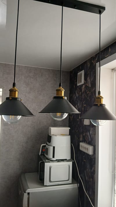 The image shows a domestic interior, likely a kitchen, in the city of Kolyvan, Russia. Three industrial-style pendant lights with black conical shades and brass accents hang from a black ceiling fixture. Each light has a clear glass bulb enclosure, revealing Edison-style bulbs. Below the lights, on a counter, sit several appliances: a white microwave oven, a white air fryer stacked on top of it, and what appears to be the top of a refrigerator. The wall behind the appliances features a dark, patterned wallpaper, contrasting with the textured gray wallpaper on the left wall. A white electrical outlet strip is visible on the patterned wall, and a window with light-colored curtains is partially visible on the right. The overall scene is well-lit, likely by natural light from the window and the artificial lighting. There are no people present in the image.