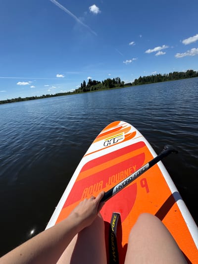 This is a first-person perspective image taken from a stand-up paddleboard (SUP) on a body of water, likely a lake or a calm river, under a bright blue sky with scattered white clouds. The foreground shows a portion of the orange and white paddleboard, with the brand "AQUA JOURNEY" and "HYDRO-FORCE" visible. A person's arm and hand are resting on the paddleboard, holding the paddle. The paddle has the "HYDRO-FORCE" branding as well. The water is dark and slightly rippled, reflecting the sunlight. In the background, a dense line of green trees forms the shoreline, with a few taller trees visible on the right side. The horizon is clearly defined between the water and the sky, with faint white contrails from an aircraft visible in the upper left quadrant of the sky. The overall scene depicts a peaceful, outdoor recreational activity on a sunny day. The specific location is identified as Lendži, Latvia, suggesting this is a natural body of water in that region.