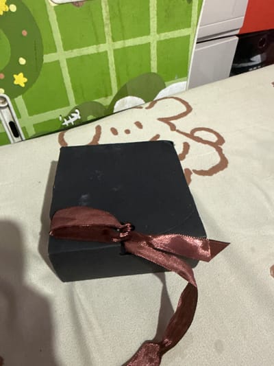 A small, black, hexagonal gift box is presented on a beige, textured surface. The box is tied with a shimmering, dark pink ribbon that forms a bow at the top and trails off to the right. The surface is decorated with scattered brown cartoonish shapes resembling flowers or clouds. To the left of the box, a green surface with a white grid pattern is visible, suggesting a mat or a notebook. This green surface has some cartoonish drawings of stars and circles. In the background, partially obscured, are what appear to be stacks of white and black electronic devices. The lighting suggests it is daytime, possibly indoors, given the lack of strong natural light. The overall impression is a neatly packaged gift, perhaps ready to be given. The image does not display any people, specific time of day, weather conditions, or overt emotional cues. There is no visible text in the image. The location context provided is Manila, Philippines.