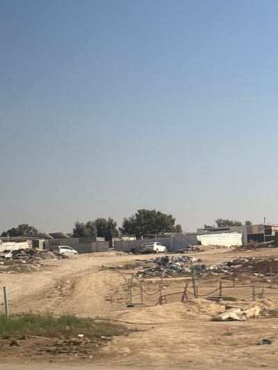 The image shows an outdoor scene with a clear, light blue sky taking up the left portion of the frame. The right side of the image depicts a dusty, unpaved road with some buildings and vegetation. The buildings appear to be under construction or in a state of disrepair, with corrugated metal sheeting visible on some of them. There are several cars, including a white one parked near some trees and debris. Piles of what looks like scrap metal and construction materials are scattered along the roadside. The overall impression is of a somewhat undeveloped or industrial area. The lighting suggests it is daytime, likely with bright sunlight, though the sky appears hazy. There is no visible text in the image.