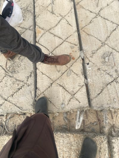 The image depicts an overhead view of a construction or demolition site in Gyellesu, Nigeria. The ground consists of a rough, light grey concrete surface featuring an irregular grid pattern of grooves and cracks. Sections of exposed steel rebar are visible, particularly along linear breaks in the concrete. Two individuals are present. One person, positioned towards the upper left, wears dark grey trousers and brown lace-up work boots; a white hard hat rests on the ground near their left foot. The second person, positioned at the bottom center, wears dark brown trousers and dark grey perforated clogs. A black pipe or cable, partially embedded in the concrete, runs vertically through the right side of the frame, with white string-like material tied around it towards the bottom. Concrete fragments and debris are scattered across the textured surface.