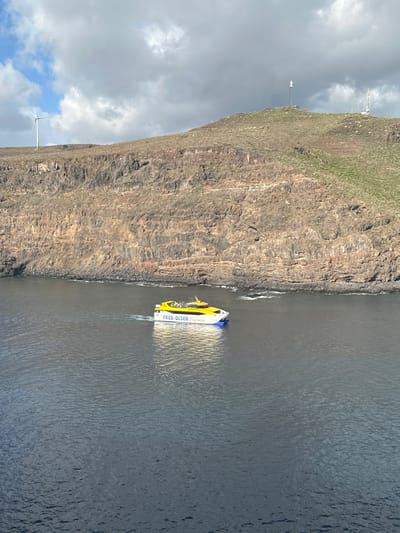 The image displays a yellow and white Fred. Olsen Express catamaran ferry sailing in dark blue waters near the volcanic coastline of San Sebastián de la Gomera, Spain. The ferry, emblazoned with "FRED. OLSEN Express," is actively moving from left to right across the frame, leaving a white wake behind it. In the background, a steep, layered cliff face of brown and reddish-brown volcanic rock rises directly from the sea, where small waves are visible at its base. Above the cliffs, the terrain ascends into a hillside covered with sparse, dry, yellowish-green vegetation. A white wind turbine with three blades is situated on a distant ridge on the upper left. On the highest point of the hill to the right, a white statue, identified as the Monumento a la Virgen de Guadalupe, is mounted on a pole, accompanied by additional communication towers. The sky is largely overcast with heavy grey clouds, though a patch of blue sky and lighter clouds is visible in the upper left quadrant. The scene is captured from an elevated perspective overlooking the water and coastline.