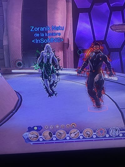 The image depicts a scene from a video game, likely a superhero-themed MMORPG such as DC Universe Online. Two avatars are visible on a futuristic, purple-hued platform. The avatar on the left is clad in a white and green costume, emitting green energy from its hands and surrounded by dark, wispy effects. Above this avatar, a name tag reads "Zoranik Nanu de la lumiere <InSoMNiO>". The avatar on the right wears a dark, armored suit with glowing red accents and is enveloped in fiery red energy. The environment features hexagonal patterns on the walls and a metallic floor. A user interface element with various circular icons and the label "L2" is visible at the bottom of the screen, indicating in-game controls or abilities. There is no information in the image or context provided that places this scene in Nantes, France.