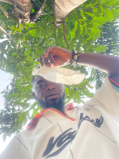 A low-angle shot depicts a person with dark skin holding a crumpled white tissue in their right hand. The person has short black hair and a beard. They are wearing a white shirt with a black pattern and an orange zipper. The person's arm is adorned with a bracelet made of black beads, interspersed with cowrie shells and beads in Rasta colors. The background features green leaves and a tree trunk, suggesting an outdoor setting. The location is Ogbe, Nigeria.