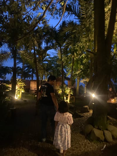 In Tamiajeng, Indonesia, a man in a black shirt embraces a girl wearing a light dress. They stand on a pebbled path near a rocky slope. Trees, some illuminated by lights, surround them. The sky appears blue through the foliage.