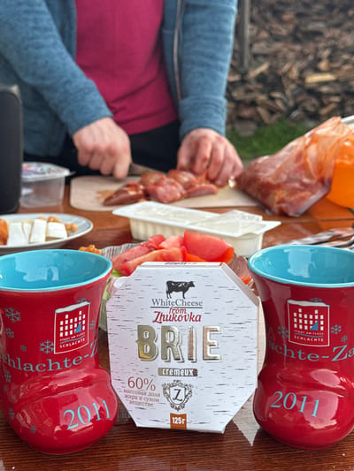 The image captures an outdoor scene with a wooden table as the primary surface. A person, partially visible from the torso down, is in the process of slicing meat on a wooden cutting board. Their attire includes a blue sweater over a pink shirt.
Scattered on the table are various food items. In the foreground, two red mugs with blue interiors are prominent, each featuring a logo and the year "2011" in blue lettering. One mug has snowflake designs. Between the mugs, a box of "BRIE" cheese, with text indicating "White Cheese from Zhukovka," is positioned. The cheese box has a stylized birch bark design and mentions "60% жира в сухом веществе" (60% fat in dry matter) and "125г" (125g).
To the left of the cheese box, a plate holds slices of cheese and nuts. Further back, a white container holds a creamy substance, possibly yogurt or sour cream, and a platter displays sliced tomatoes and lettuce. A large bag of red meat is visible on the right side of the table, partially obscuring an orange object. In the background, a stack of firewood suggests a rustic or rural setting. The overall ambiance is informal, possibly a picnic or outdoor meal preparation. There is no information in the image that specifies a location within the city of Maksimovo, Russia.