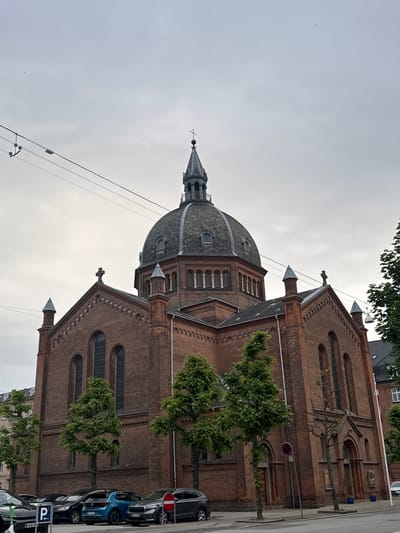 The image shows a large, red brick building with a domed roof and spires, likely a church or a similar public building. The architecture is detailed, with arched windows and decorative brickwork. The building is surrounded by trees, some of which are in full leaf, suggesting it is spring or summer. Several cars are parked along the street in front of the building, indicating an urban setting. One of the cars is blue, and another is black. There are also power lines and poles visible, typical of a street scene. The sky is overcast and a light grey, suggesting cloudy weather. The time of day appears to be daytime, but the diffused light indicates it is not sunny. The overall scene is quiet and still, with no people visible. There are some signs visible on the street, including a "no parking" sign with a red circle and a horizontal line, and a blue parking sign with a "P" symbol. The location context is Frederiksberg, Denmark, which aligns with the architecture and the presence of parked cars.