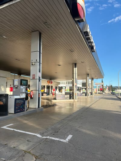 The image shows a Repsol gas station in Rubí, Spain, during daylight. Gas pump number 7 is in the foreground. The facade of the gas station building is visible, displaying the Repsol logo. Across the street are various store signs, including "El Rai de les Pintures" and "LOW COST R 654 350 www.trasteros." A vehicle is visible in the distance on the road.