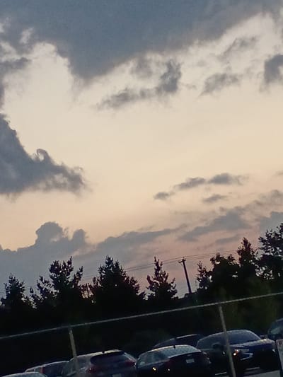 The image shows a cloudy sky with the sun setting over a line of silhouetted trees and a chain-link fence in the foreground. Several cars are parked behind the fence, some with their headlights on. The sky is a mix of dark grey and pale orange clouds, with some thinner, wispy clouds scattered throughout. There are no people or animals visible in the image. The scene appears to be an outdoor parking lot, possibly at dusk. The image does not provide any specific information about the location within the city of Lexington, United States of America.