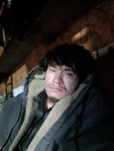 A close-up, slightly upward-angled image depicts one male individual indoors. The subject, an adult male, has dark, wavy hair and facial hair consisting of a mustache and goatee. He is wearing a dark-colored, hooded jacket with a visible beige sherpa lining, open over a dark inner layer. His gaze is directed toward the camera with a neutral expression. The background is dimly lit and consists primarily of rough, light-colored wooden paneling or plywood, with darker, indistinct elements occupying the upper portion. Shadows are pronounced due to the low-light conditions. No specific identifiers for the city of Ozark, United States, are discernible within the visual content of the image.