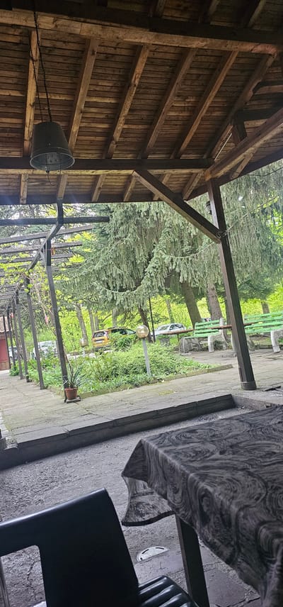 The image shows an outdoor scene in Sveti Konstantin, Bulgaria, partially covered by a wooden structure. The wooden structure features visible beams and planking. A metal bucket hangs from a chain attached to the wooden beams. Beyond the wooden structure, there's a garden or park area with trees, a park bench, a street lamp, and some vegetation. A table and chair, partially visible, are near the edge of a paved area next to the wooden structure. Cars are visible further into the background. The image is taken from a slightly elevated perspective.