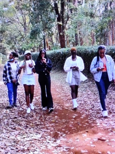 The image depicts five individuals walking on a dirt path covered with scattered dry leaves in an outdoor, vegetated area of Dodoma, Tanzania. A dense green hedge forms a boundary behind the path, backed by tall trees with full green foliage.
From left to right:
1. A female subject wears a black and white plaid shirt over a light top, blue jeans, and a light blue face mask. Her gaze is directed towards the right.
2. A female subject, wearing a white baseball cap, white top, light blue ripped denim shorts, and white sneakers, smiles forward while holding a light-colored bottle.
3. A female subject, dressed in a black button-up top and black flared pants, holds a small cylindrical object to her mouth.
4. A female subject, with short light-colored hair, wearing an oversized white shirt, light shorts, and white socks, is focused on a smartphone held in both hands.
5. A female subject, wearing a light blue denim jacket over an orange-red top, dark jeans, and eyeglasses, is also focused on a smartphone held in both hands.
The group is in active transit, with two subjects engaged with mobile devices and one engaging with a handheld object at her mouth.