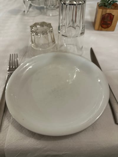 The image displays an overhead view of a dining table set for a meal. A large, round, white dinner plate occupies the center-right of the frame, appearing empty and with a slight sheen reflecting ambient light. A silver knife is positioned above the plate, angled upwards and to the left, while a silver fork is partially visible below the plate, angled downwards and to the left.

To the left of the plate, two clear glass tumblers are visible. The one closer to the plate is a tall, ribbed glass, while another, similar glass is partially cropped on the far left. A third, shorter, clear ribbed glass is located below the main plate, also partially visible.

In the upper-left corner of the image, a small, light brown wooden box is present. A small, green, artificial plant or decorative element sprouts from its top. The box features a label with a dark red background. Clearly visible text on the label reads "MEZEDOPWΛIO", identifying it as a Greek meze restaurant. Below this text, there is a graphic resembling a bunch of grapes or a wine bottle. Further text beneath the graphic is partially visible and appears to read "TO ΞHPH".

The table is covered with a white tablecloth, which shows some natural wrinkles and folds. The overall setting suggests a casual dining environment, possibly in a restaurant or tavern in Greece, awaiting guests or food. No people are present in the image, and there is no specific activity taking place beyond the table being set.