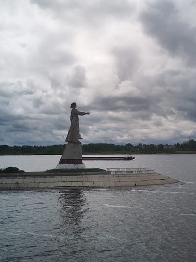 The image depicts the Mother Volga statue in Rybinsk, Russia, situated on an artificial island in the Volga River. The statue is of a woman in flowing robes with her arm outstretched. Behind the statue is the river, with a barge visible, and trees lining the distant shore. The sky is overcast with heavy, dark clouds. The statue is placed on a concrete base, surrounded by a small grassy area and a white balustrade. The river occupies a significant portion of the foreground.