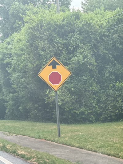 The image shows a yellow diamond-shaped traffic sign with a left-pointing arrow and a red octagon shape on it. The sign is mounted on a gray metal pole. Lush green foliage fills the background. To the right, there is a strip of grass and a gray concrete sidewalk. The image was taken in Mountain Park, United States of America.