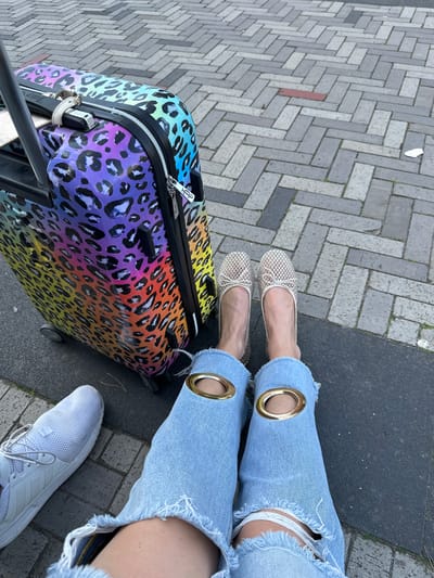 The photograph depicts a person standing on a paved surface, likely a sidewalk, near what appears to be a street in Dronten, Netherlands. The paving is composed of interlocking gray bricks, with a single red brick visible. The person is wearing ripped light blue jeans with decorative gold rings at the thigh, a pair of beige ballet flats, and a white athletic shoe. Next to the person is a colorful suitcase featuring a leopard print design in a gradient of colors including yellow, orange, red, purple, and blue. The suitcase has a black handle and zipper, as well as a silver luggage tag. A strip of dark gray asphalt runs along the edge of the paved area, suggesting the edge of the street or another surface. The overall setting is urban, and the photograph appears to have been taken outdoors during daylight.