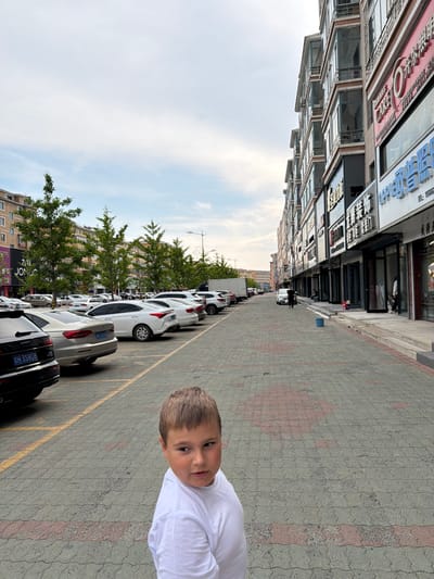 The image shows a street scene in Hunchun, China. In the foreground, a young boy wearing a white t-shirt looks to his right. The background features a row of multi-story buildings with shops on the ground floor. Cars are parked along the street, and trees line the sidewalk. The sky is overcast.