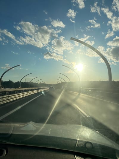 The image shows a view from inside a vehicle traveling on a highway. The highway is elevated with protective barriers on the sides. Arch-shaped streetlights line the highway, extending over the lanes. The sun is bright and low in the sky, creating lens flare and illuminating the clouds. There are other vehicles on the highway in the distance. The surrounding environment appears to be a wooded area. The image is taken in Beloostrov, Russia, which is part of the Vyborgsky District of Leningrad Oblast.