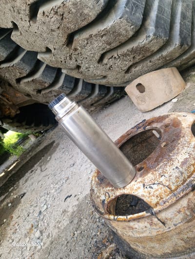 An upright, metallic thermos with a black cap rests inside a rusted, circular metal object with multiple holes. To the left of the thermos, a large, heavily treaded tire is visible. Behind the tire, a rectangular, light-colored object with a hole in its center lies on the ground. The ground surface consists of gravel and dirt. In the background, green foliage is partially visible. The scene appears to be outdoors, possibly in an industrial or construction area. The "Galaxy S22+" watermark is present in the bottom left corner of the image.