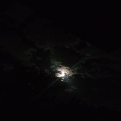 The image captures a nighttime scene in Ashtarak, Armenia, where the moon is partially obscured by dark clouds. The moon's light, however, shines through breaks in the clouds, creating a radiant glow. The surrounding sky is predominantly black, indicating the darkness of night. The clouds themselves are textured and appear somewhat wispy, with some illuminated by the moonlight and others remaining dark and shadowed. The overall impression is one of a moody and atmospheric night sky, with the moon being the central focal point. There are no people, animals, or man-made structures clearly visible in the frame.