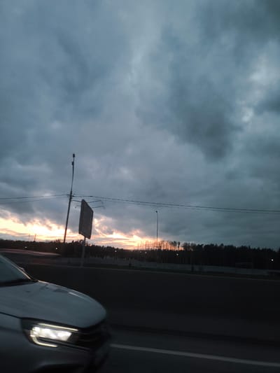 The image depicts a dusk scene in Novoivanovskoe, Russia, viewed from a moving vehicle. The sky is dominated by heavy, dark clouds, with a band of orange and yellow light visible on the horizon, suggesting a setting sun. In the foreground, the illuminated headlight of a silver car is visible. A road runs horizontally across the lower part of the image, alongside which there is a roadside barrier. Beyond the barrier, there is a line of trees silhouetted against the fading light. Two vertical light poles and a road sign are also present. Power lines extend across the frame.