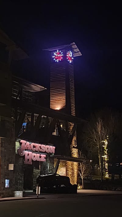The image displays a nighttime scene of a building in Teton Village, United States of America. A large, illuminated sign with pink-red lettering spelling "Jackson Hole" is mounted on the stone-clad facade of the lower structure. Above and to the right, a tall, slender clock tower constructed of horizontal wooden planks rises. The clock tower features two visible clock faces, each outlined with blue neon lighting and containing red, snowflake-like internal illumination against a white background. A single upward-facing yellow light illuminates the lower section of the wooden tower. An elevated dark-beamed wooden walkway or bridge connects sections of the building, passing in front of the clock tower's base. The lower left portion of the building exhibits stone masonry. In the immediate foreground, a dark paved surface is visible. To the right of the entrance, an irregularly shaped, dark object covered in white material, possibly snow or ice, rests on the ground. A small, light-colored vertical marker is positioned on the paved surface nearby. To the far right, a tree wrapped with numerous warm white string lights stands among other bare trees against the dark night sky. No individuals are present in the scene.
