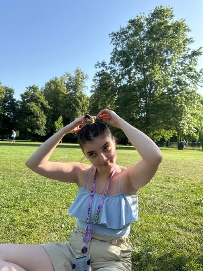 A young woman with dark hair pulled into a bun sits on a grassy lawn in a park, facing the camera. She is wearing a light blue ruffled tank top and beige shorts. A lanyard with multiple tags hangs around her neck. Her hands are raised, adjusting her hair. In the background, several large trees with green foliage are visible, and a clear blue sky fills the upper portion of the image. The grass is green and appears well-maintained, with some dappled sunlight. In the distance, a few other people are faintly visible walking on the lawn. This scene is consistent with a public park in the city of Epsom and Ewell, United Kingdom, such as Nonsuch Park or Ewell Court House grounds, which are known for their open green spaces and mature trees.