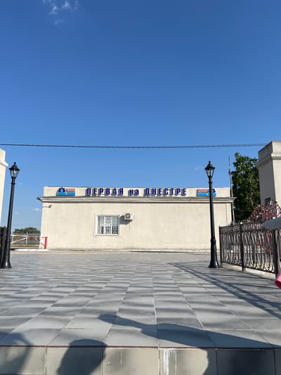 A low-angle, full shot captures a bright, sunny day with a clear blue sky in Korzhevo, Moldova. In the center of the frame, a one-story, light-colored building is visible. Blue Cyrillic lettering that reads "ПЕРВАЯ НА ДНЕСТРЕ" (First on the Dniester) is prominently displayed across the top of the building's facade. To the left of the lettering, there is a logo with the number "1955" and a graphic of water. To the right, there is another logo with the number "2025" and a graphic of water. A white-framed window with ornate metal bars is centered on the building's facade, and an air conditioning unit is mounted above it. To the left and right of the building, two black lampposts with ornate designs stand tall. The ground in front of the building is paved with large, gray square tiles arranged in a checkerboard pattern, casting long shadows. In the foreground, shadows of two people are cast on the tiled ground, suggesting their presence just outside the frame. A black metal railing with circular designs runs along the right side of the tiled area, partially obscuring a red structure in the background. A white gate with red and white striped poles is visible to the left of the building, behind a short fence. Trees can be seen behind the fence line. The overall scene appears to be an outdoor public space, possibly a plaza or walkway, leading up to the building.