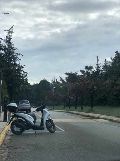 The image shows a white Honda scooter parked on the right side of a paved road in Agia Paraskevi, Greece. The road is bordered by a sidewalk and a row of trees on the right. A grey car is parked further down the road in the background. The sky is overcast and the weather is cloudy. The image was taken at a location with the coordinates 37.998 latitude, 23.819 longitude. A green electric vehicle charging station can be seen in the foreground on the left side of the road. The charging station appears to be unused. There are no people in the image.