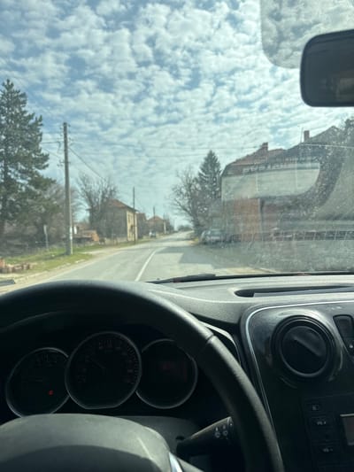 The image presents a first-person view from inside a vehicle, looking forward through a dirty windshield onto a street in Dolna Beshovitsa, Bulgaria. The lower left of the frame is occupied by a black steering wheel and a partial view of the instrument cluster, which displays a speedometer scaled to 210 km/h, a tachometer at zero, and a digital odometer reading "136974 km". An oil pressure warning light is illuminated. The dashboard's right side reveals an air vent and part of the infotainment system.
Beyond the windshield, a paved asphalt road extends into the distance, curving slightly right. Buildings with tiled roofs and weathered facades line both sides of the road, interspersed with bare deciduous trees and tall evergreen pines. Utility poles with power lines run parallel to the road on the left. A light-colored car is parked on the right side of the road further down. Reflections on the dirty windshield include a partially visible rearview mirror in the upper right and a large billboard advertisement with unreadable text and a partially visible phone number "0700 70 300". The sky above is mostly clear, featuring numerous scattered, white cumulus clouds.