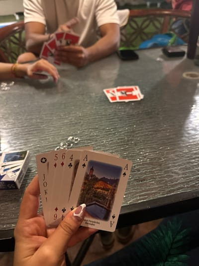 A tabletop scene with playing cards scattered. A person on the left holds a hand of red-suited cards. A hand on the right holds a fan of cards, including an Ace of Spades with a building depicted and printed text reading "Garmisch-Partenkirchen, Mohrenplatz." A deck of cards is visible in the lower-right corner. The table surface is textured, and the surroundings appear to be indoors. There is no indication of the location being Playa de Chachalacas, Mexico.