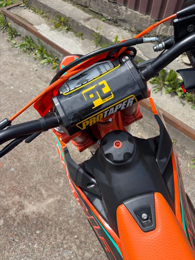 The image shows a close-up of the handlebars and fuel tank of an orange and black dirt bike. The bike is parked outdoors on a paved surface with some greenery and concrete steps in the background. The handlebars feature a black pad with yellow "PRO TAPER" branding. The fuel tank is black with a textured orange cap. There are no people visible in the image. The scene appears to be a typical outdoor setting, possibly a street or a yard.