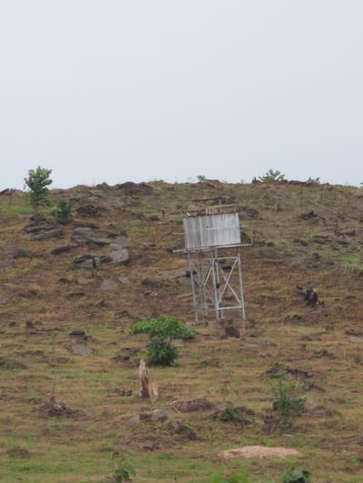 A medium shot of a rugged hillside in Kaninkon, Nigeria, with sparse vegetation of brown and green hues. The hillside is punctuated with rocks and small trees. At the summit of the hill stands a metal frame structure with a white, flat rectangular object or screen attached to one side. The sky is overcast and light gray. The image is rotated counterclockwise.