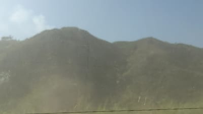 A hazy landscape shows rolling, vegetation-covered hills under a light blue sky. The foreground is blurred, appearing as if viewed through a dirty window. There are several scratches in the lower right part of the window. The scene lacks sharp details, suggesting a dusty or foggy atmosphere.