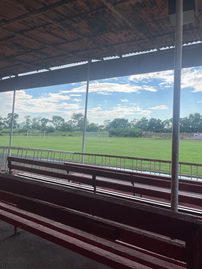 The image captures a ground-level view from beneath a covered spectator stand at a sports field in Chirpan, Bulgaria. In the immediate foreground, multiple rows of dark reddish-brown wooden bleachers are arranged on a dark, textured ground surface. A light-colored metal safety railing runs horizontally across the scene, separating the seating area from the field. Two prominent, light-colored vertical support poles, likely metallic, anchor the roof structure; one is positioned on the right side of the frame, and another is slightly left of center, partially behind the railing. Above, the underside of a corrugated roof, exhibiting signs of wear and age, with visible support beams, spans the top of the frame.
Beyond the railing, a verdant green grass football (soccer) field extends into the midground. A white metal goal frame is positioned on the left side of the field, while a second goal, equipped with a net, is visible on the right. A dense line of green trees and natural vegetation forms the backdrop to the field. On the far right, partially obscured by trees, sections of dilapidated tiered seating or stands, featuring faded blue and red seats, are discernible. The sky above is a clear blue, dotted with scattered white cumulus clouds. No people or animals are present in the scene.