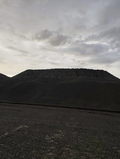 The image depicts a vast, dark landscape dominated by a large, conical pile of dark material, likely coal or slag, under a cloudy sky. The material forms undulating hills and a prominent, tiered summit. A dark, linear feature, possibly a road or railway track, runs horizontally across the foreground. The sky is overcast with soft, diffused light. The environment appears industrial or post-industrial, devoid of any visible plant life or human presence. The photograph was taken in Shal'skii, Russia.