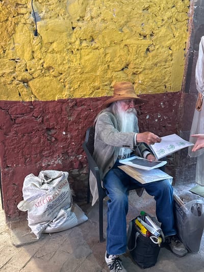 The image shows an elderly man with a long white beard sitting in a gray plastic chair against a two-toned wall (yellow and red) in San Miguel de Allende, Mexico. He wears a straw hat, a gray blazer, and blue jeans. He is holding several items: some papers with the word "BIO" printed on them and two white, framed photographs or documents. A person is standing over him, possibly handing him the papers. Next to the man on the ground are several bags and items: A white bag labeled "AZUCAR ESTANDAR", a black backpack, a shopping bag with the word "BACCO" on it, and a collection of miscellaneous items. The setting is likely a street or alleyway.