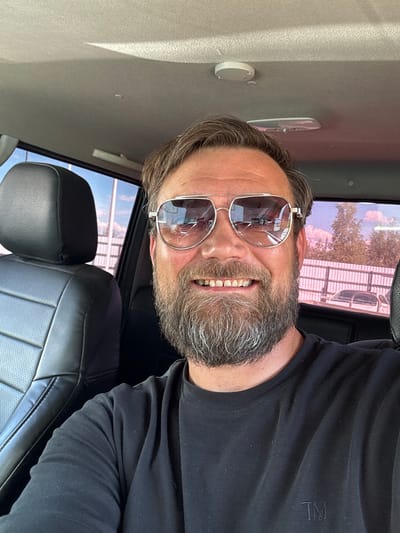 A bearded man wearing sunglasses and a black t-shirt smiles for a selfie inside a vehicle. The interior of the vehicle is visible, including a black leather seat to the left and the interior roof. Outside the vehicle, a pink fence and trees are visible through the tinted windows. The man appears to be in Parma, Russia, as indicated by the context provided, though no specific landmarks confirm this location within the image itself.