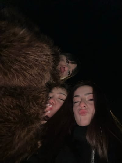 Three women pose closely together for a selfie taken from a low angle. The woman on the left has brown hair with bangs, peering out from behind a large, brown, fur-like garment that the other two women are also nestled in. The woman in the upper right has long, dark hair and puckered lips. The woman nestled beside her, on the lower right, has similar dark hair and is also puckering her lips, with her hand visible near her face, showcasing long, white fingernails. The background is dark.