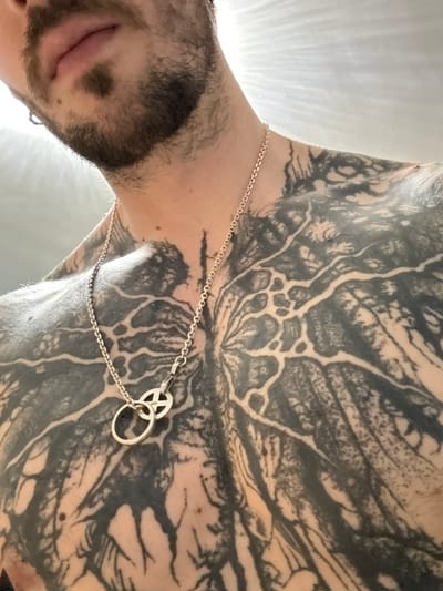 A close-up, eye-level, over-the-shoulder shot shows a person's chest and lower face, tattooed extensively with black ink in a pattern resembling tree roots or veins. A silver chain necklace with two circular pendants is visible on the chest. The background is a bright, out-of-focus white, suggesting a well-lit indoor environment. The individual has a light-colored beard and a small hoop earring in their left ear. The image is set in Putuo District, China.