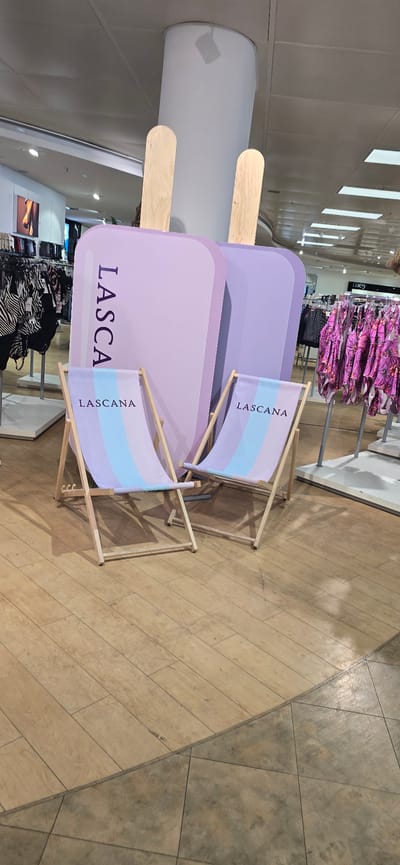 This image shows a retail store display featuring a large, purple, popsicle-shaped prop with "LASCANA" written vertically in a lighter shade of purple on one side. Two beach chairs with the "LASCANA" logo are placed in front of the prop. The beach chairs have fabric seats with colorful pastel stripes. In the background, clothing racks are visible, with mannequins displaying clothing, including swimwear with floral patterns and zebra-print items. The floor is made of light-colored wooden planks. The scene appears to be set up in a store, possibly a department store or a clothing boutique. This image was taken in Gütersloh, Germany, specifically within the Hugendubel bookstore and cafe, located at Kirchstraße 11-13.