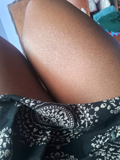 The image depicts part of a person's body, specifically their thigh, and a portion of a dark dress with a floral pattern. The skin has a smooth texture and golden hue, with visible hair follicles. The dress features an intricate white design on a black background, resembling a paisley or mandala pattern. The overall composition is a close-up shot, focusing on the textures and patterns of the skin and fabric.